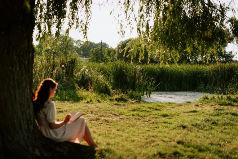 A woman reading a book under a willow tree beside a pond on a calm summer evening
