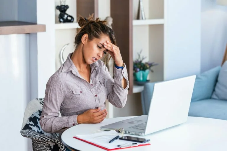 A woman at a desk with her hand on her forehead, looking doubtful in front of her laptop