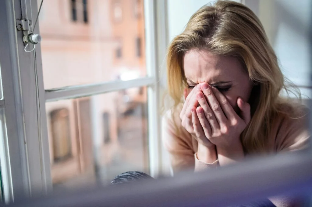 A woman by a window with her hands over her face, feeling low