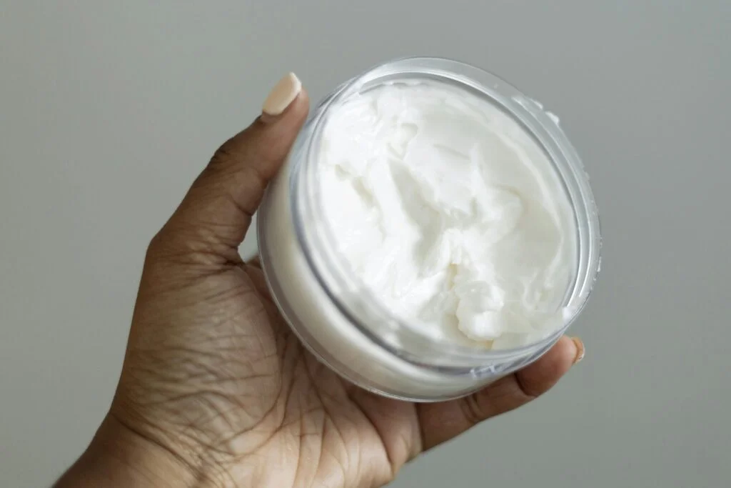 A hand holding an open jar of white body cream