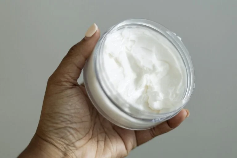 A hand holding an open jar of white body cream