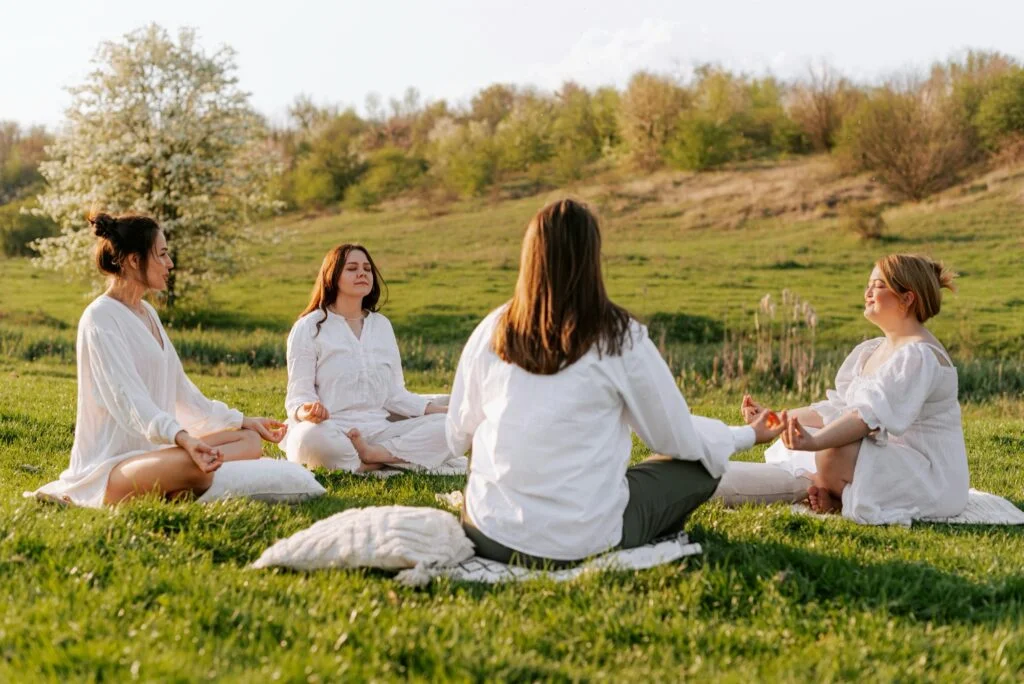 A group of women in white sitting cross-legged on grass meditating with their eyes closed