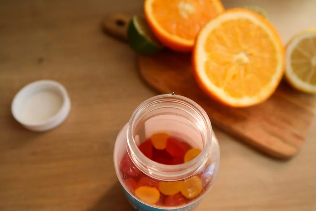 Open jar of fruit-flavoured supplement gummies beside sliced oranges on a kitchen table