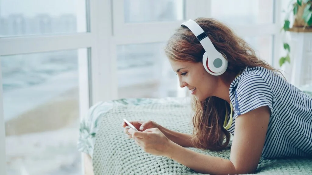 A smiling woman lying on a bed wearing headphones and choosing music on her phone