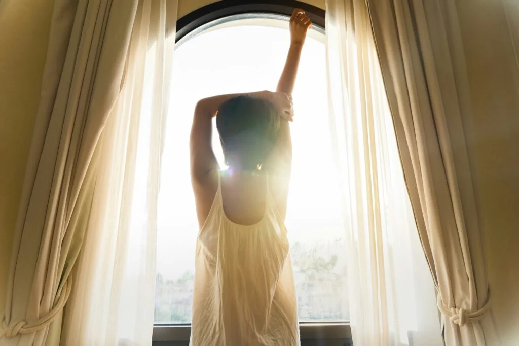 A woman stretching in front of a bright window as morning sunlight streams in