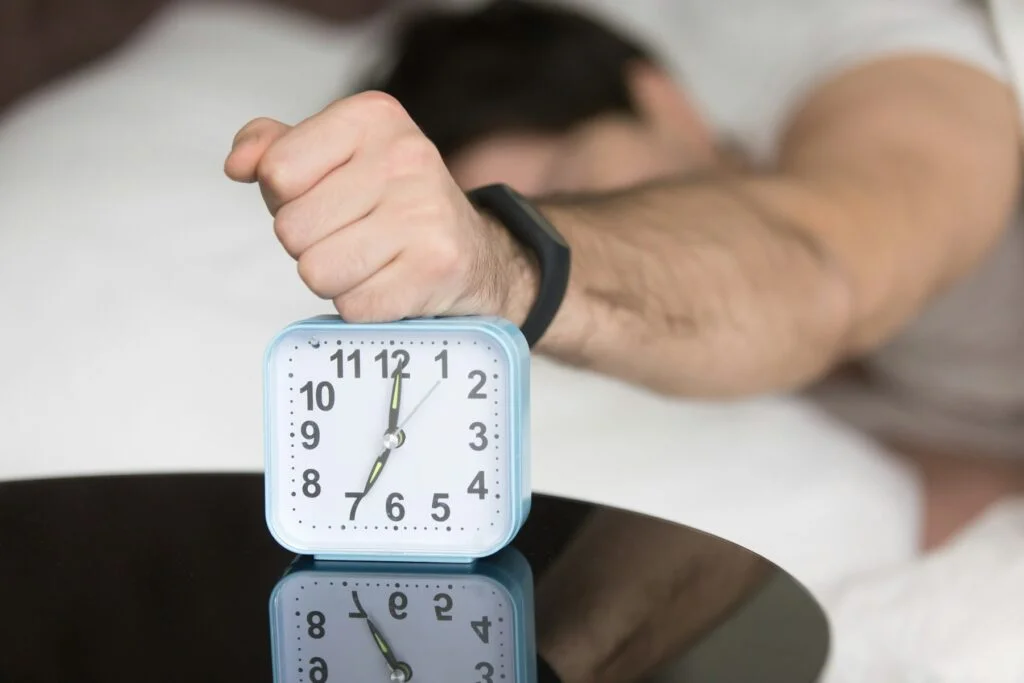 Hand pressing the snooze button on an alarm clock beside the bed