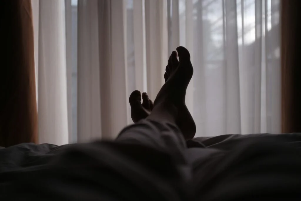 Silhouette of a person's bare feet raised in bed against light curtains at night