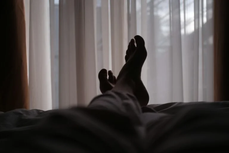 Silhouette of a person's bare feet raised in bed against light curtains at night