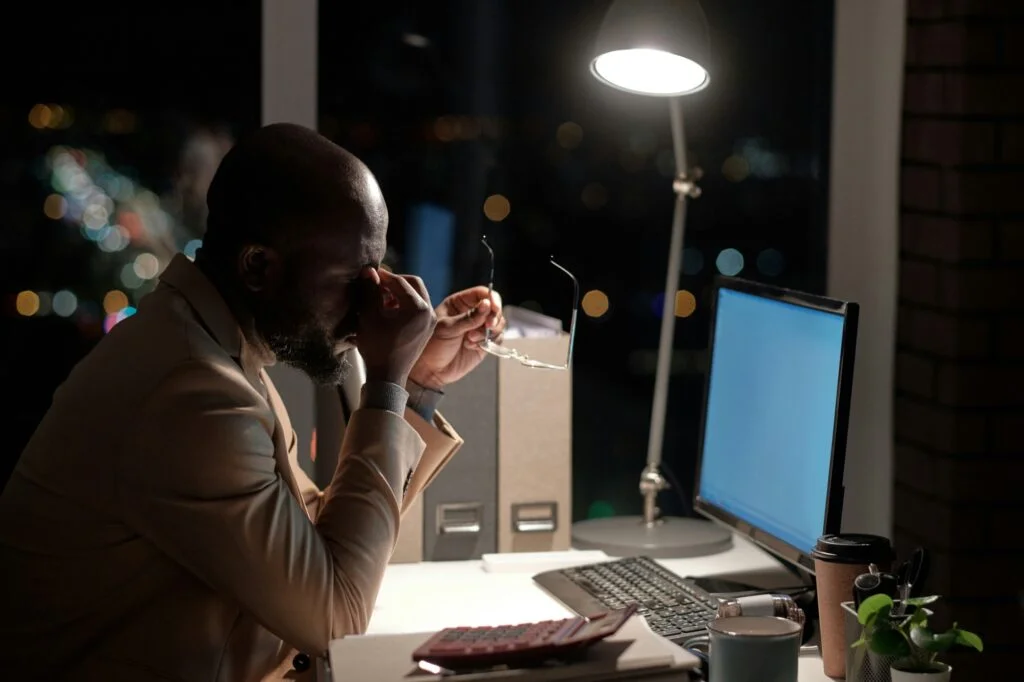 An exhausted man rubbing his eyes at his desk late at night