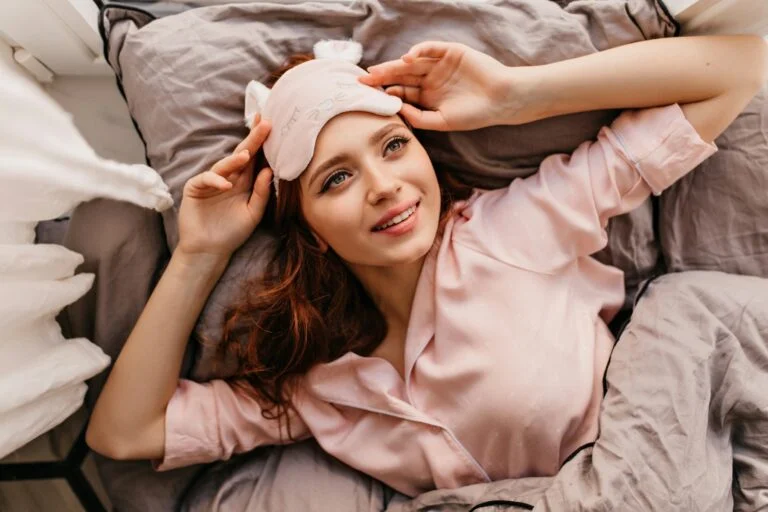 A smiling woman in pink pyjamas lying in bed with a sleep mask pushed up on her head