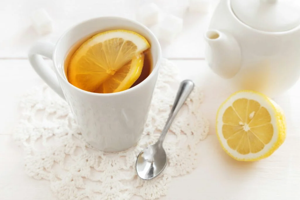 A white mug of hot tea with a slice of lemon beside a teapot