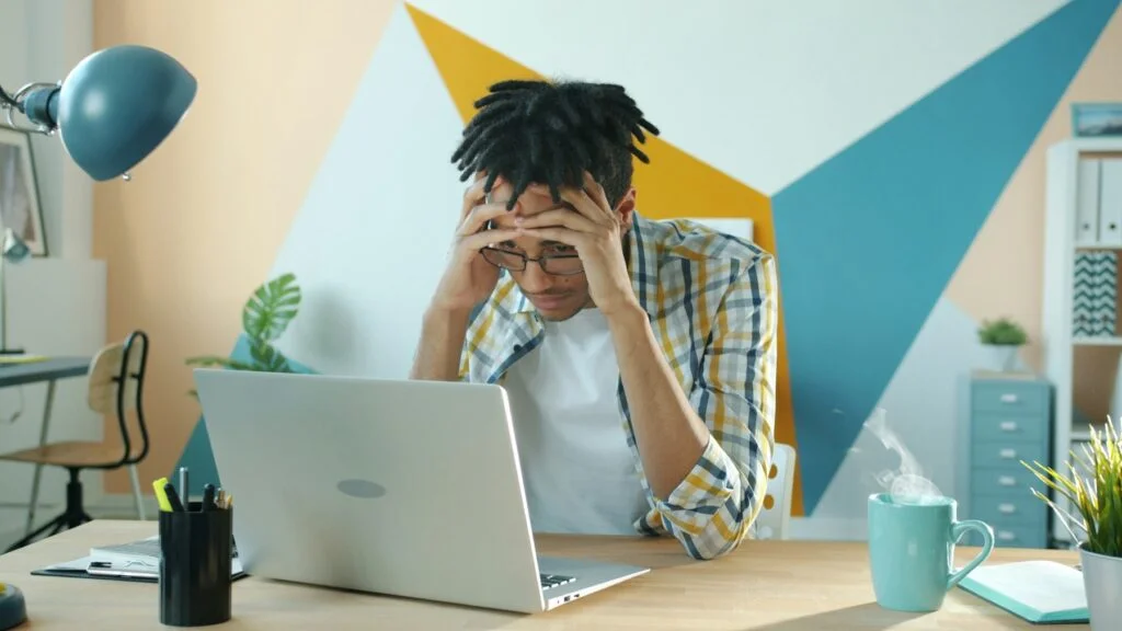 A man at his desk holding his head in his hands in front of a laptop