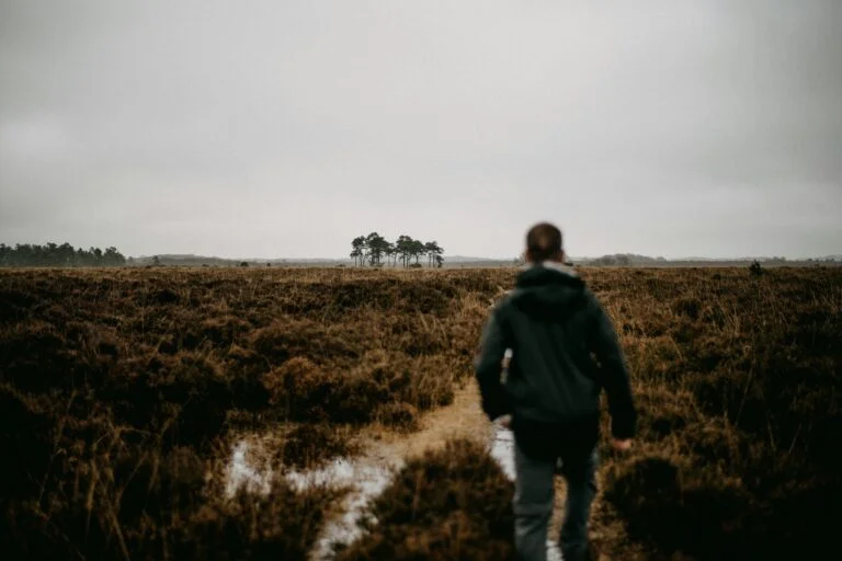 A man in a dark jacket walking along a path across open heathland on a grey day