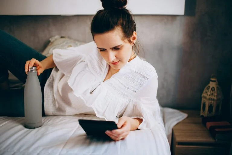 A woman resting on a bed with a drink and a tablet