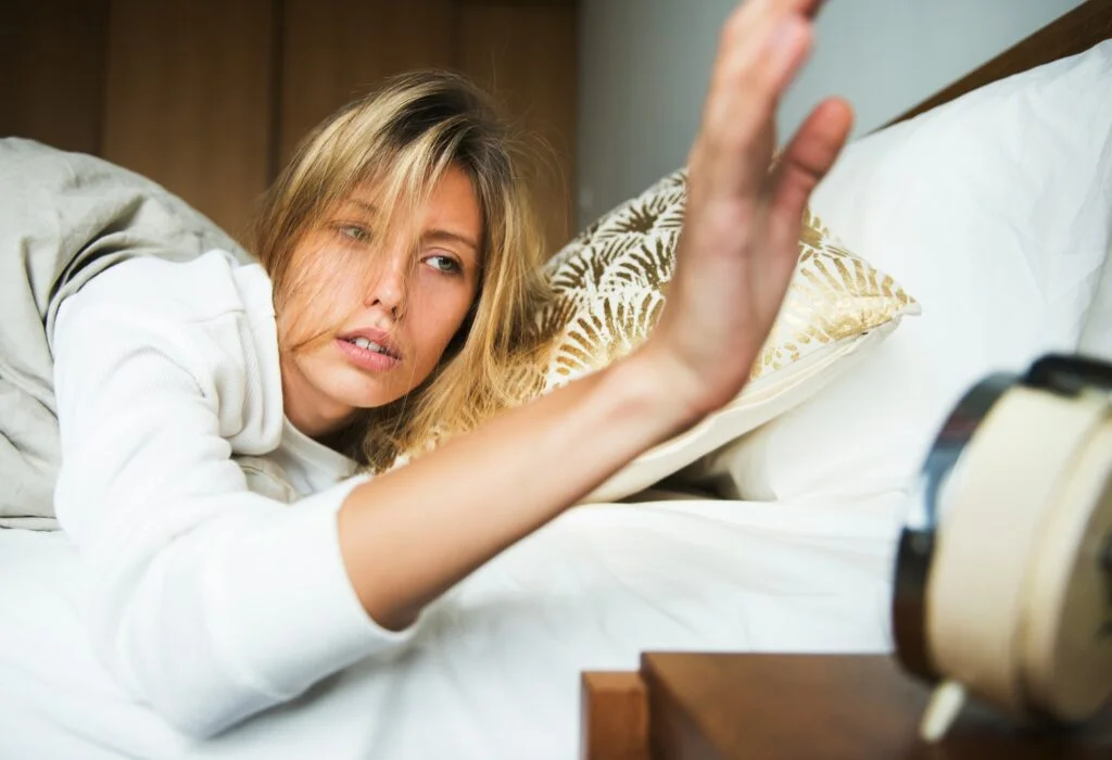 A tired woman in bed reaching out to switch off her alarm clock