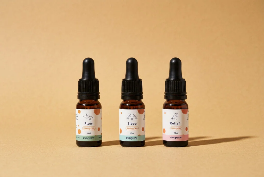 Three Evopure CBD oil bottles, Flow, Sleep and Relief, standing in a row on a warm sand background