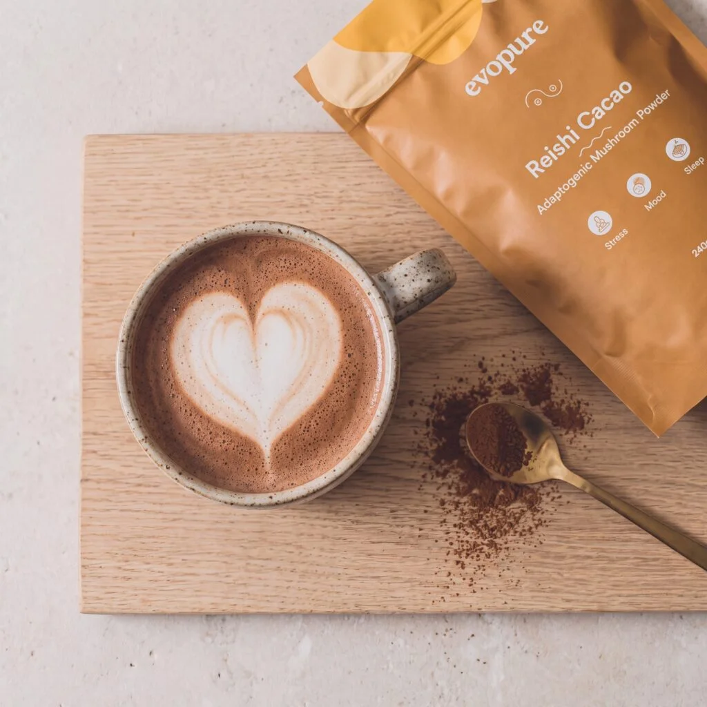 Mug of reishi hot chocolate with a heart latte beside a pouch of Evopure Reishi Cacao on a wooden board