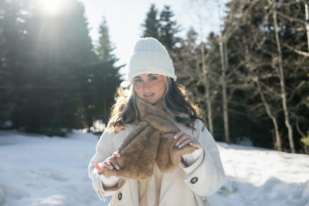 A smiling woman in a white hat and faux fur scarf outdoors on a sunny, snowy winter day