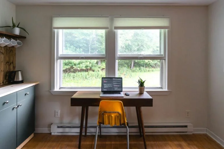 A simple home office desk with a laptop and plant in front of a window